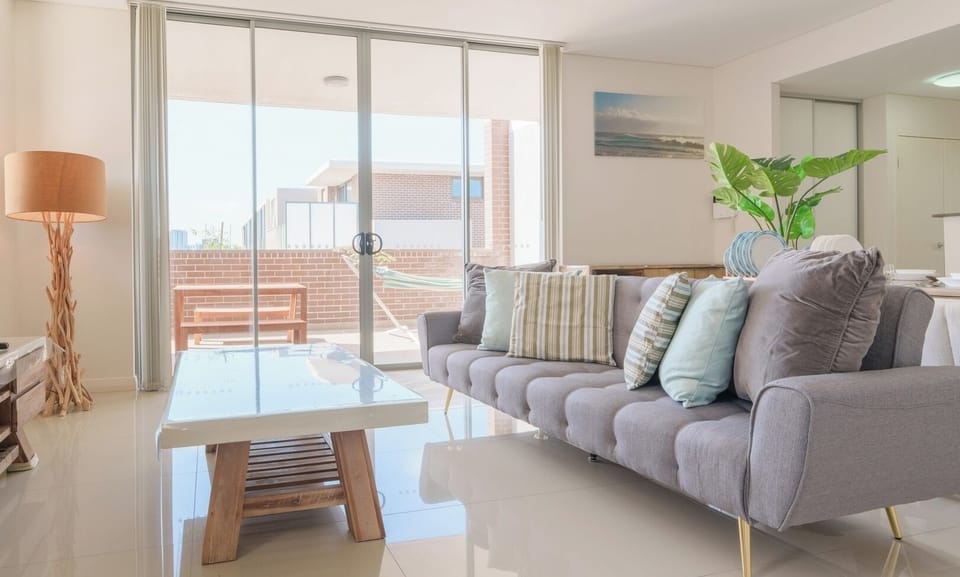 A spacious living area highlighting a long grey sofa, a low wooden coffee table, and wide sliding glass doors that open to an outdoor balcony.