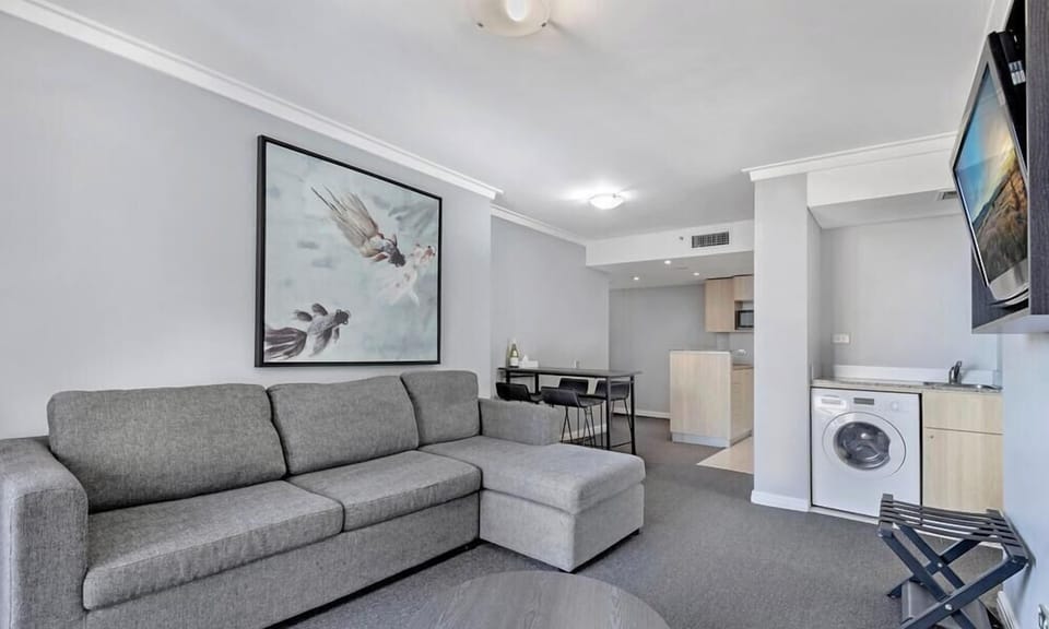 An open-plan living space showcasing a grey L-shaped sectional sofa, neutral carpets, a decorative wall painting