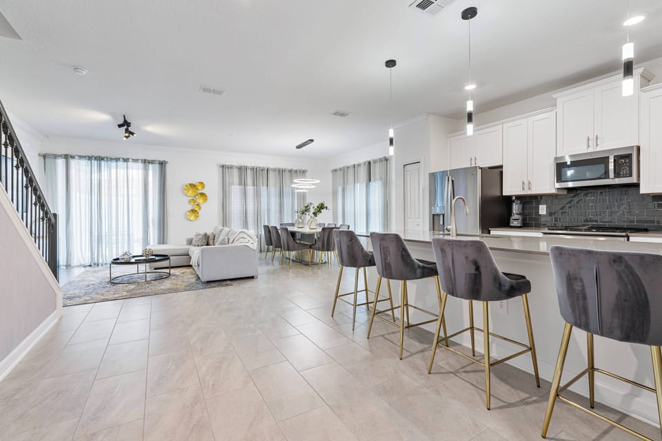 Experience a bright and spacious living area featuring sleek gray barstools around the kitchen island. Sunlight floods in through large windows, enhancing the modern furniture and tasteful decor for a relaxing atmosphere.