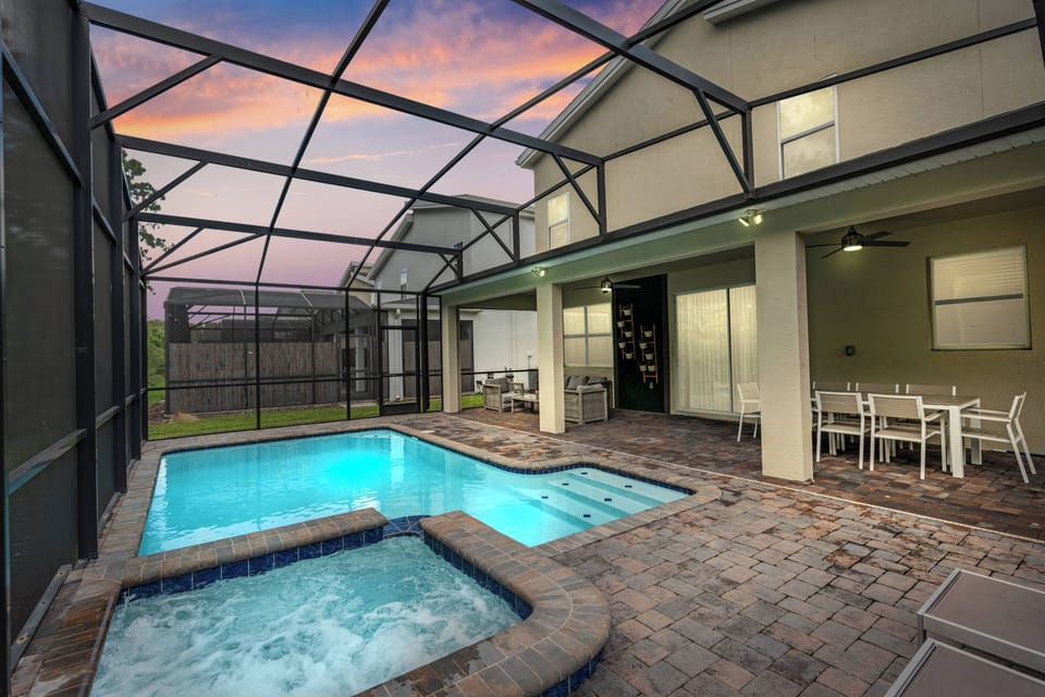 Step outside to find a beautifully paved patio featuring a sparkling pool and a relaxing hot tub, all framed by a sleek screen enclosure. The twilight sky casts a warm glow, creating a perfect ambiance for evening gatherings.