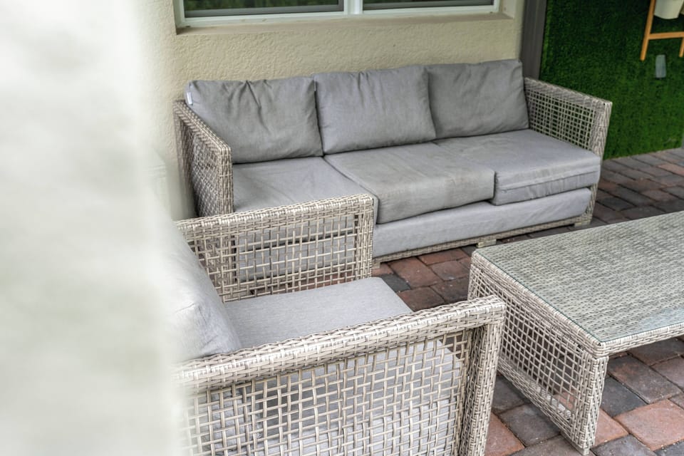Relax on the cozy light gray sofa amid stylish woven furniture, enjoying the gentle breeze. The rustic stone patio is ideal for casual gatherings, while the greenery outside adds a refreshing touch of nature to your space.
