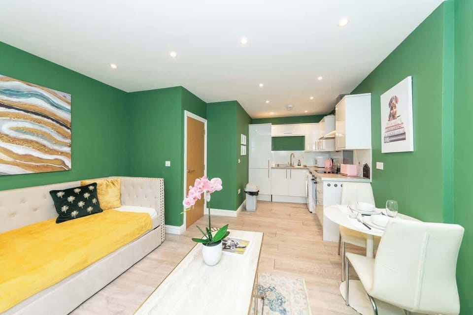A modern living room with green walls, a cream sofa with yellow and black cushions, a large abstract painting, a window with a white blind, a coffee table with a plant, and light wood flooring and a fully furnished kitchen for cooking just like home.