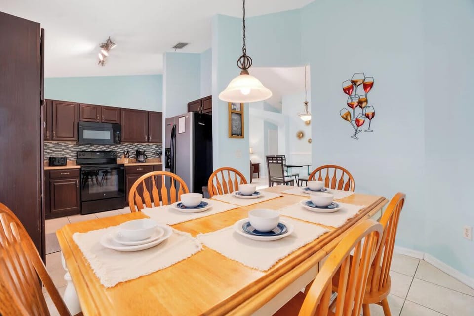 Dining area with wooden table set for six and a view of the kitchen with dark cabinetry.