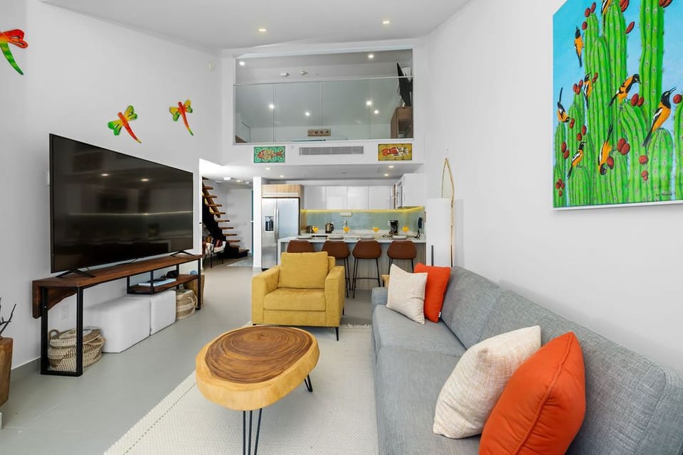 Step into your vibrant, open-concept retreat where colorful art, a cozy gray sofa, and a sunny yellow chair invite you to unwind while a fully equipped kitchen and lofted space await your every need.