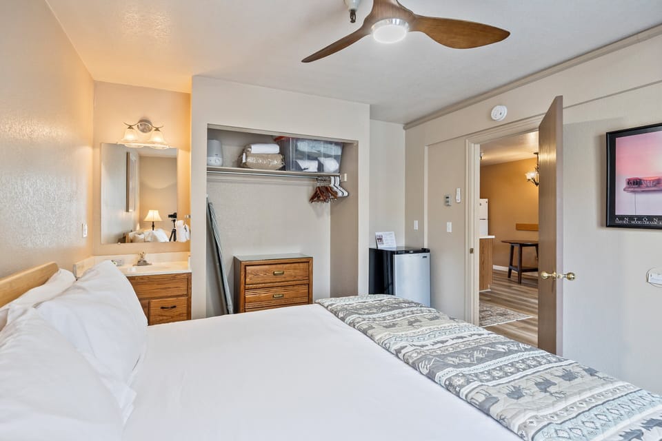 Comfortable king bedroom featuring a ceiling fan, open closet with storage, in-room vanity sink, and mini fridge. Conveniently located next to the bathroom and living area for an easy, functional layout.