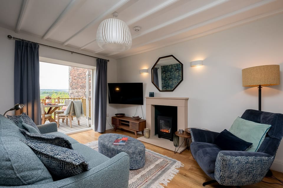 Cosy living room with sofa, armchair, fireplace, wall mounted TV and open doors leading to a balcony with outdoor seating and countryside views