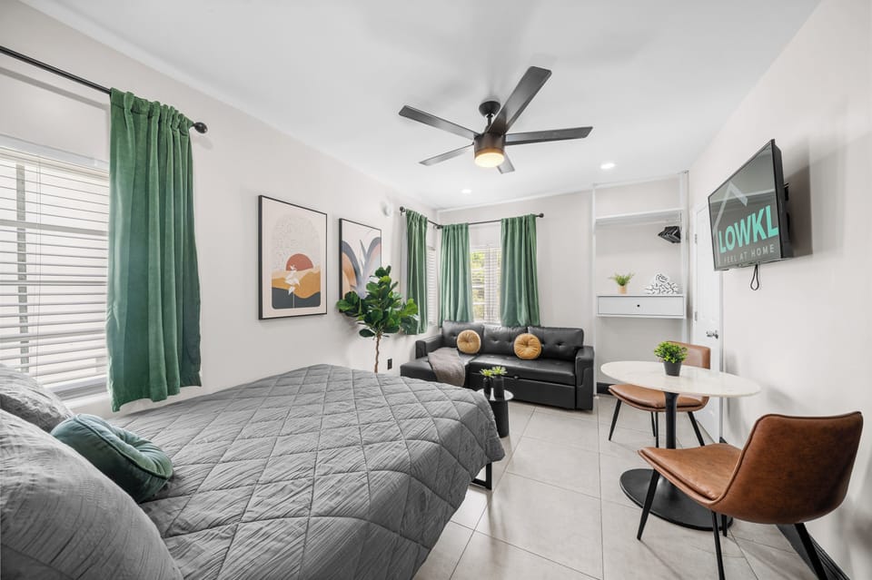 This inviting studio combines comfort and style, featuring a queen-size bed, a cozy leather sofa, and a dining area for two. The space is enhanced by vibrant wall art, natural light from windows, and soft greenery, creating a relaxing atmosphere.
