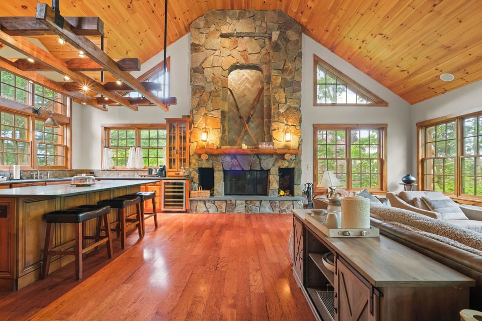 Expansive open-concept living area with a dramatic stone fireplace, vaulted wooden ceilings, and a cozy seating area. The adjacent gourmet kitchen features a large island, perfect for entertaining in this warm, rustic setting.