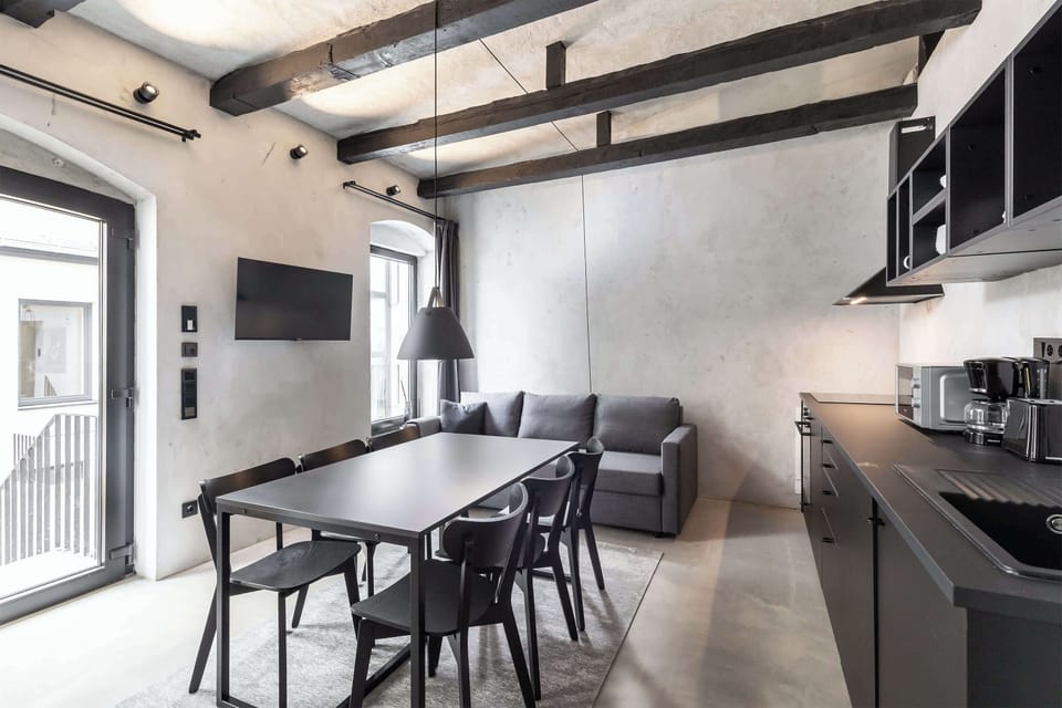 A spacious living area featuring a modern grey sofa, large windows, a wall-mounted TV, and a generous dining table. Clean architectural lines and raw textures create a refined industrial ambiance.