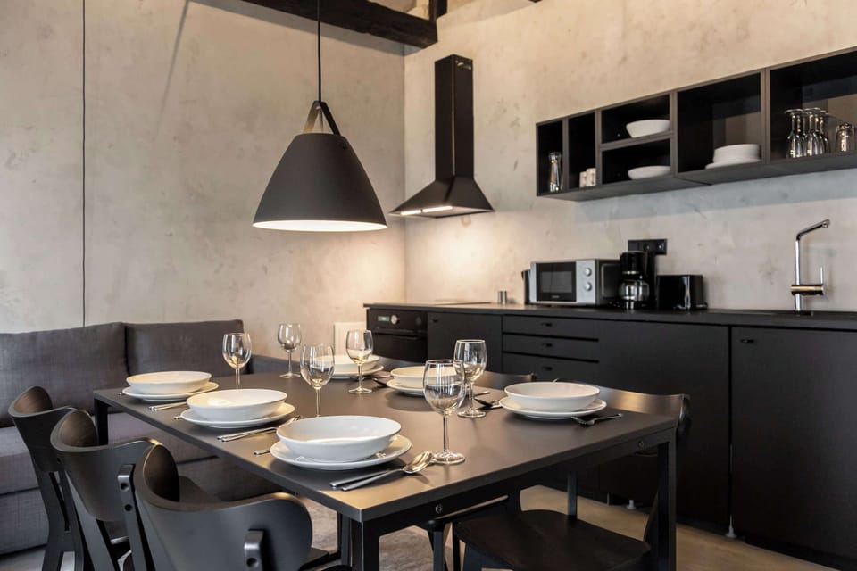A stylish dining setup with tableware for six beneath a sculptural pendant lamp. The black kitchen and textured concrete walls provide an elegant, modern backdrop.