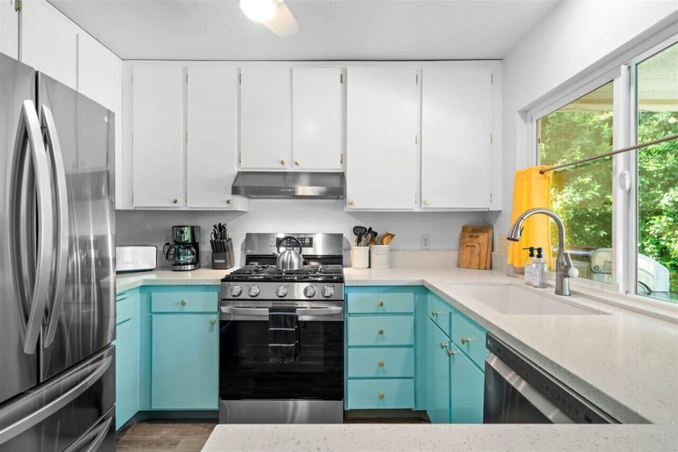 Bright, Cheerful & Fully Equipped! ☀️🍽️ This vibrant kitchen combines retro-chic turquoise cabinets with modern stainless steel appliances, offering both charm and functionality. A sunlit window overlooks lush greenery, making meal prep feel like a dream.