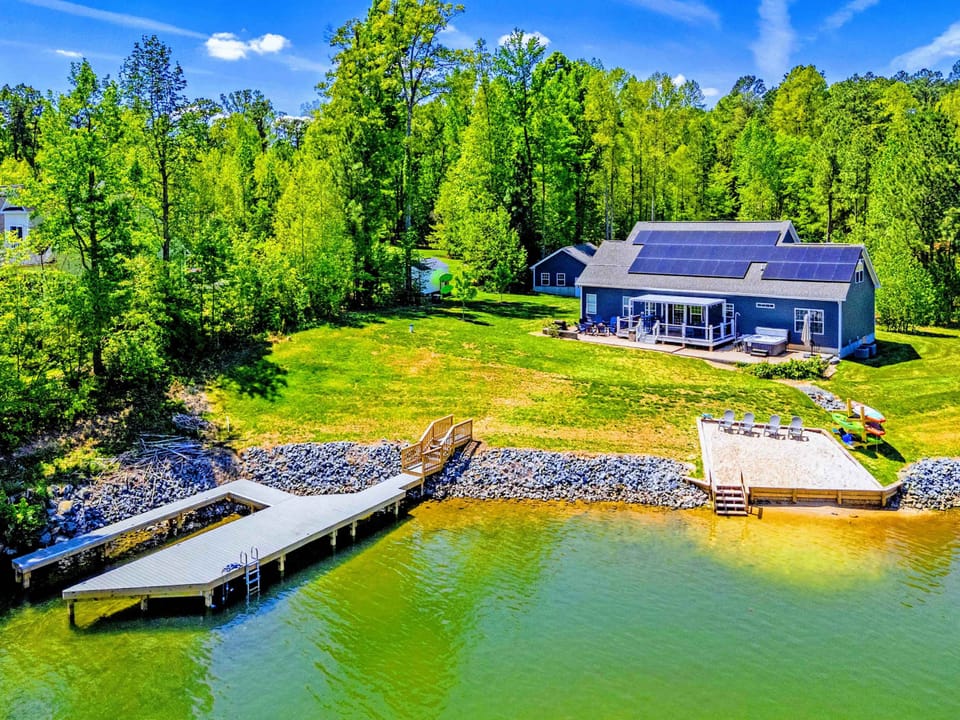 Aerial - The full lakefront view: blue craftsman home, sloped lawn, sandy launch beach, and your private dock.