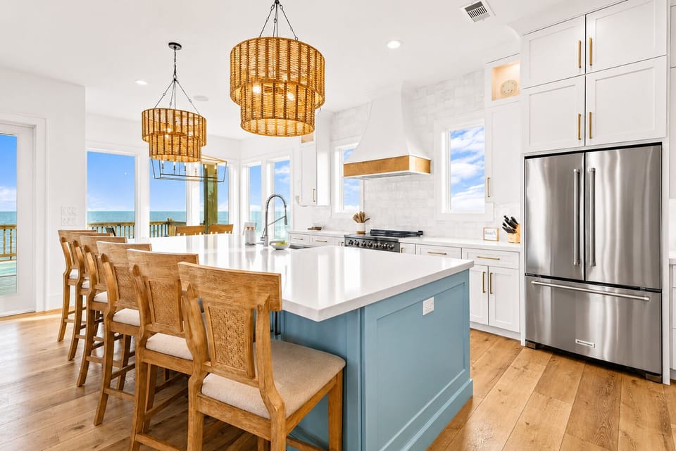 Bright kitchen with blue island, quartz-style counters, bar seating, stainless steel refrigerator, range, white cabinetry and large Gulf-facing windows.