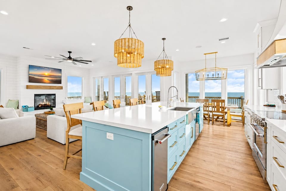Bright open-concept kitchen and living room with large island, white countertops, coastal blue cabinetry, pendant lighting and abundant windows overlooking the Gulf.