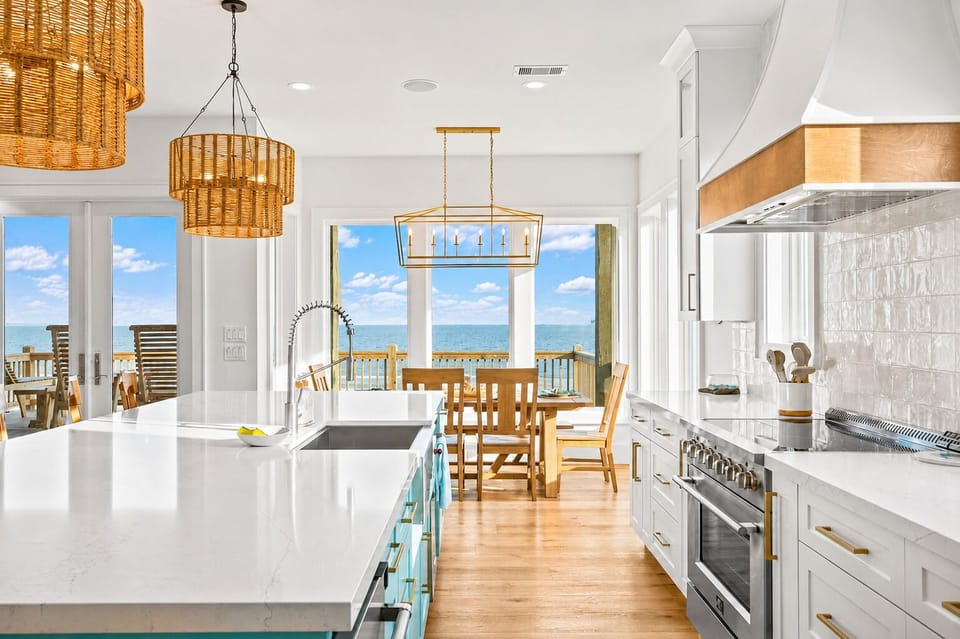 Bright kitchen featuring stainless steel range, vent hood, large island, farmhouse-style sink, pendant lighting and Gulf views through expansive windows.