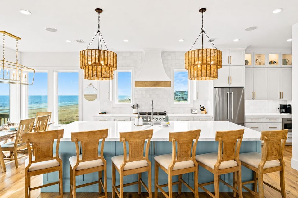 Spacious beachfront kitchen featuring a long blue island, multiple bar stools, statement pendant lights, white cabinetry and stainless steel refrigerator.