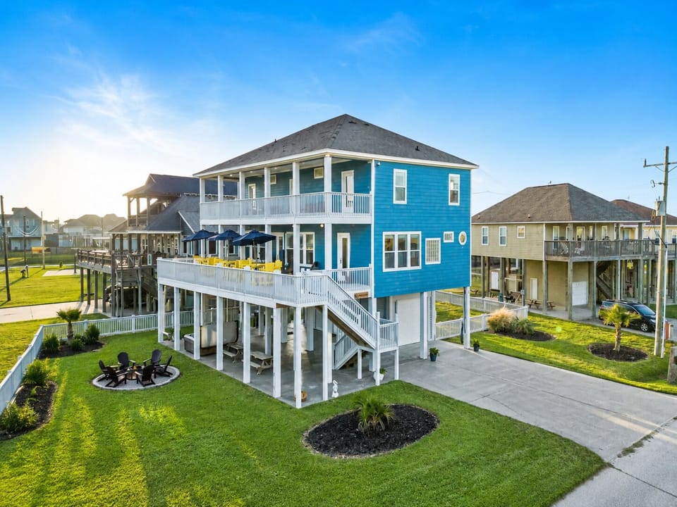 Elevated Galveston beach house exterior with wraparound deck, large yard, covered parking, and sunset coastal lighting.