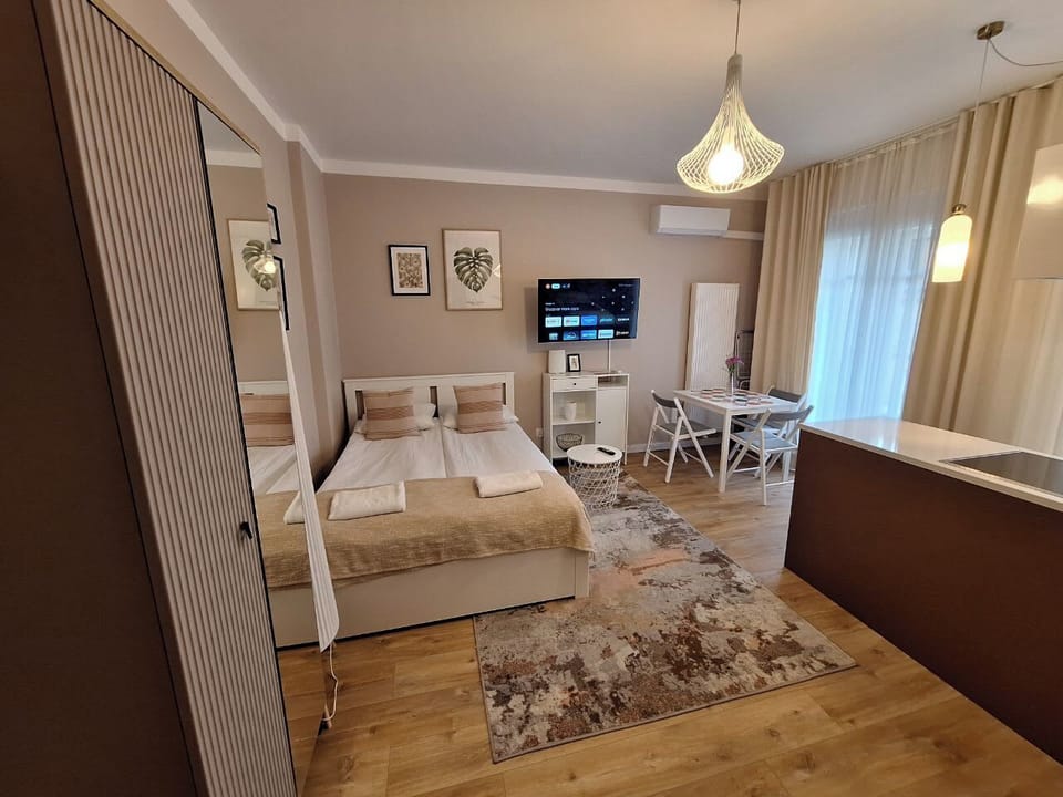 A wall-mounted TV positioned opposite the bed provides convenient entertainment for guests staying in the apartment. The bright interior and light-colored furniture make the space feel modern and inviting.