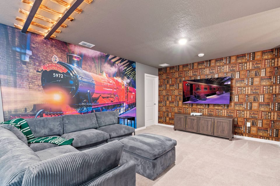 This vibrant entertainment room features a plush gray sectional couch, perfect for movie nights. The striking mural of a classic train adds a whimsical touch, while warm wood accents on the walls create an inviting atmosphere.