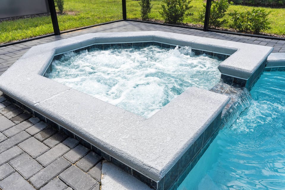 Unwind in your private oasis with a spacious hot tub, surrounded by elegant gray stonework and inviting blue water. The lush greenery outside the screened enclosure adds to the serene ambiance for a truly peaceful retreat.