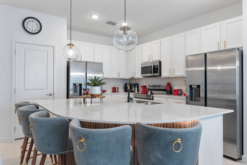 This modern kitchen features stainless steel appliances, a spacious island with plush blue seating, and crisp white cabinetry, all enhanced by stylish pendant lighting, making it a welcoming space for your culinary adventures.