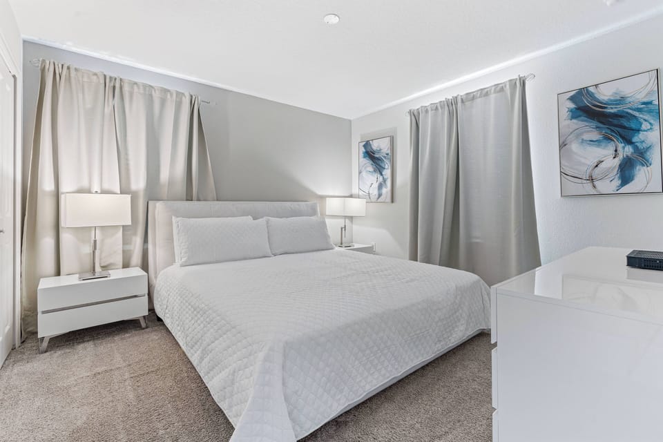 This cozy bedroom boasts a plush light-hued bed with soft linens, elegant nightstands, and warm-glowing lamps. The calming blue and gray decor sets a tranquil atmosphere, ideal for restful nights and peaceful stays.