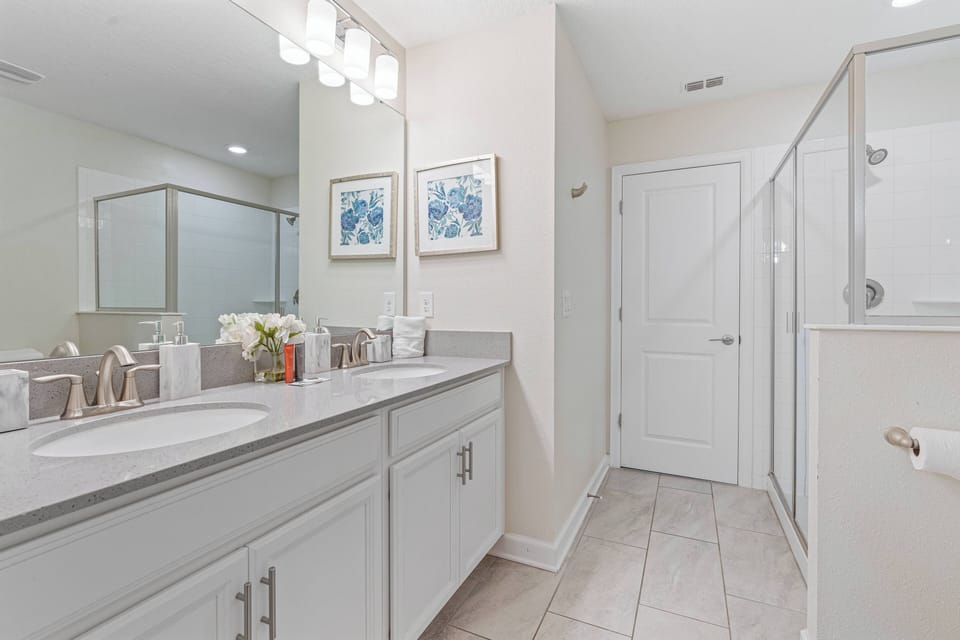 Experience a bright, airy bathroom with a spacious double vanity and sleek gray countertops, enhanced by elegant light fixtures. Fresh decor adorns the walls, and a modern glass shower adds a luxurious touch to your stay.