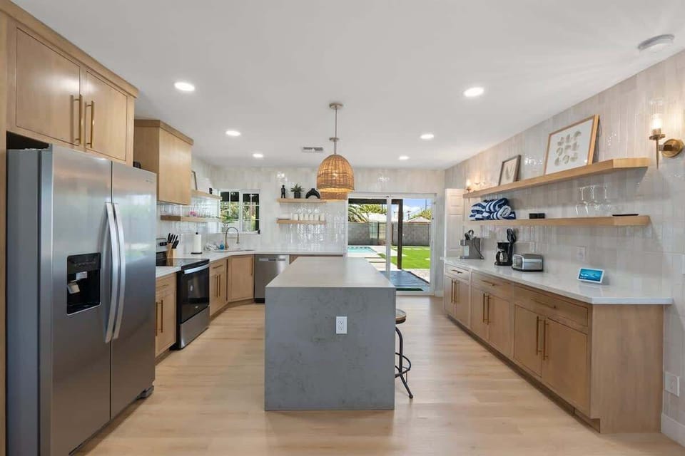 Cook and chat around the big kitchen island—stainless fridge, warm wood cabinetry, open shelves, and pendant lights that keep everything bright while you prep meals.
