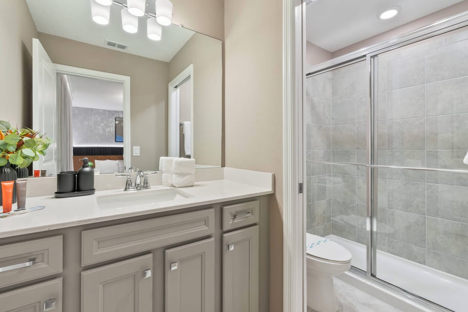 Experience a stylish bathroom with sleek gray cabinetry and a spacious vanity featuring a pristine white countertop. Enjoy the glass-enclosed shower and soft lighting that create a warm and inviting ambiance for your relaxation.