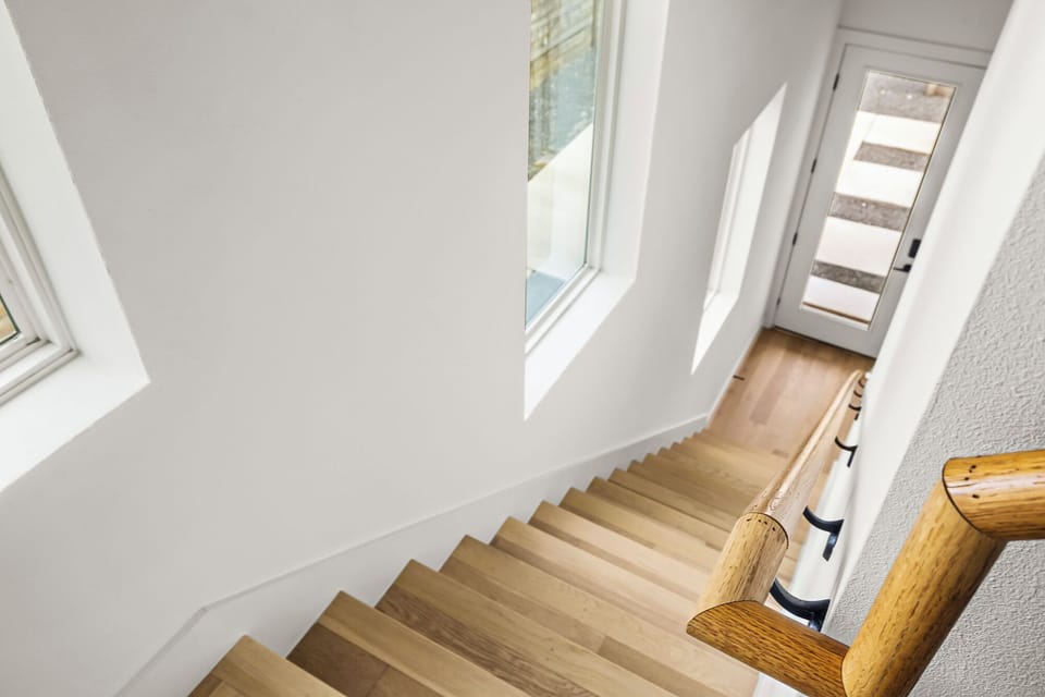 A modern staircase with light wooden steps and a matching wooden handrail. The walls are painted white, creating a clean and bright atmosphere. 