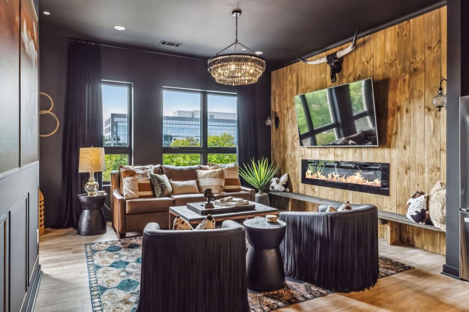 Western-themed living area with a rustic, wood-paneled accent wall featuring a mounted TV and an electric fireplace beneath it. The room is furnished with a leather sofa, black leather chairs with fringe detailing.
