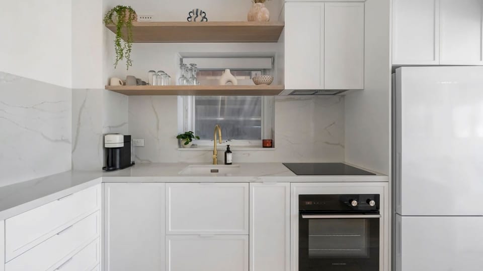 Step into this impeccably designed kitchen, where sleek white cabinetry meets warm wood shelving and elegant gold accents to create the perfect blend of modern style and functionality.