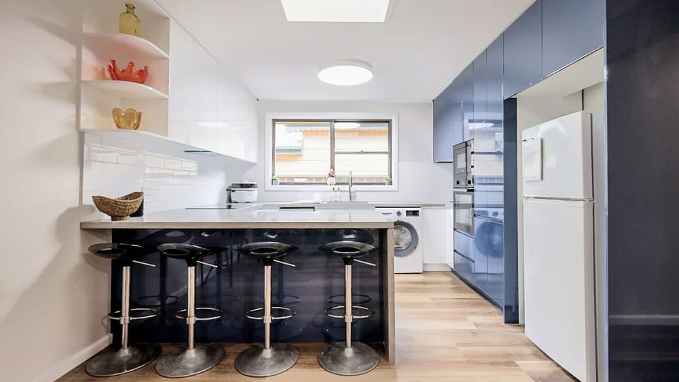 Bold design meets daily functionality. Step into a striking, modern kitchen featuring high-gloss deep blue cabinetry, crisp white accents, and a spacious breakfast bar complete with sleek black bar stools