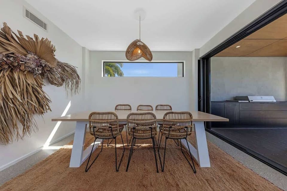 Inviting dining area features a sleek wooden table with woven chairs, a stylish pendant light, and a beautiful decorative wall piece, creating a perfect spot for meals with a view of the outdoor BBQ area.