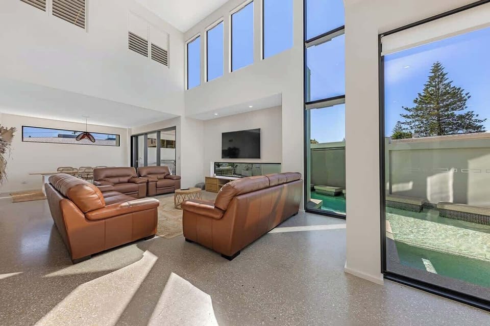 Spacious living room features comfortable leather seating, a large TV, and bright windows that open up to the outdoor pool area, creating a seamless flow between indoor and outdoor spaces.









