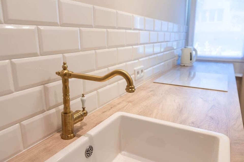 A detailed view of a brass faucet and white sink by a window. Natural light and a tiled wall elevate the kitchen design.