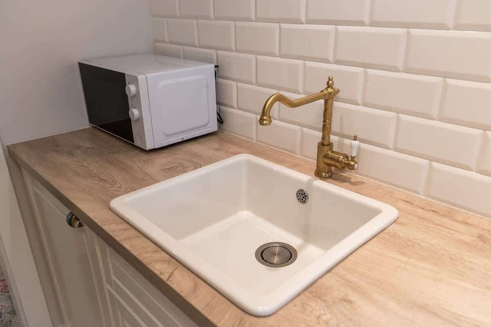 A modern kitchenette with a white sink and wooden countertop. A microwave and brass faucet complete the compact setup.