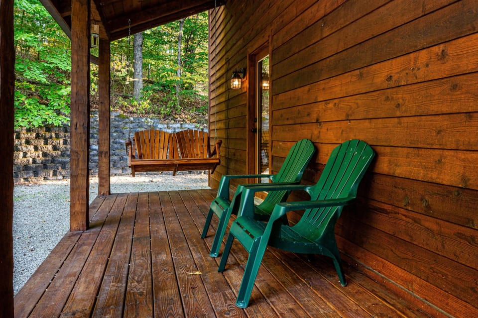 Covered wooden porch with green Adirondack chairs, a hanging wood bench, and wooded hillside backdrop.