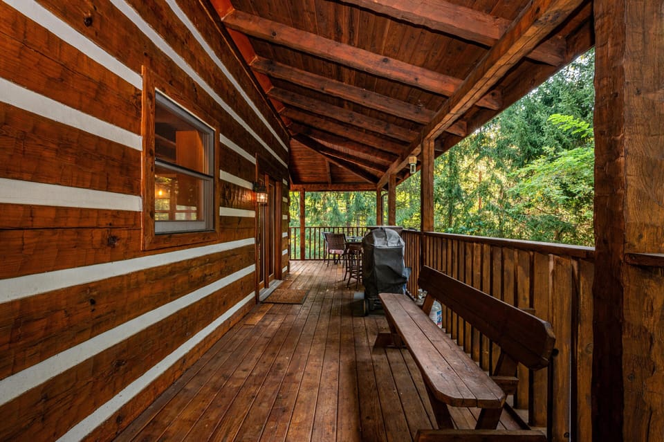 Long covered porch with polished wood planks, solid bench seating, and a charcoal grill near railing overlooking trees.