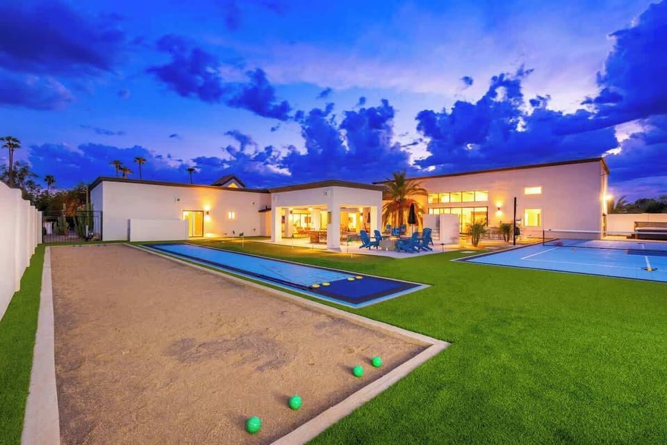 A modern desert retreat featuring bocce ball courts and sleek lap pools under evening skies, framed by manicured lawns and palm trees beyond the white stucco architecture.