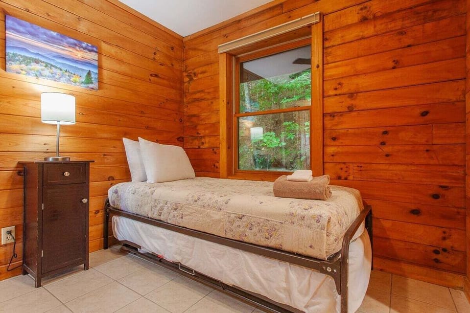 Bedroom with a twin bed, bedside table, and large window overlooking wooded surroundings. Rustic lodging near Bryson City and Great Smoky Mountains attractions.