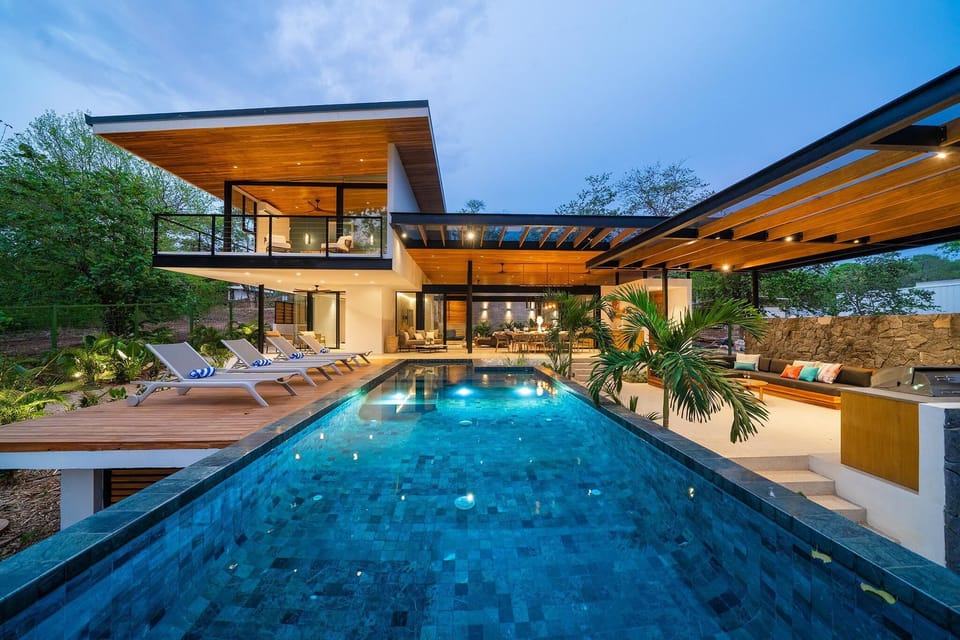 Infinity pool and view of the home, outdoor kitchen, dining, and back side of the home just after sunset