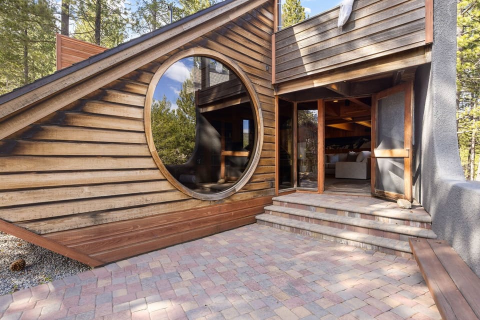 A uniquely designed architectural exterior featuring distinctive circular windows and warm wood siding, nestled among towering pines. The welcoming brick-paved entrance sets the tone for your forest retreat.