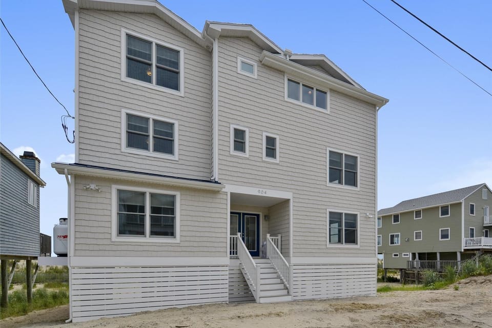 The exterior façade of a spacious multi-level beach house with coastal-style siding, steps from sandy shores and natural dune grasses.