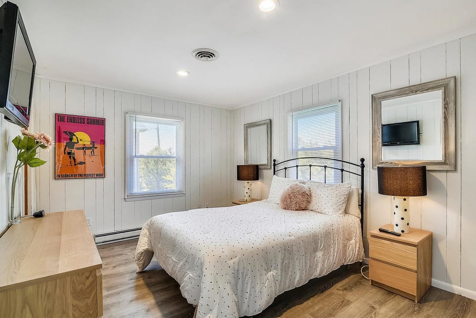 Your bright coastal bedroom features a comfy bed, polished hardwood floors, and crisp white shiplap walls. Cheerful pops of color make this space feel fresh and inviting for restful nights.