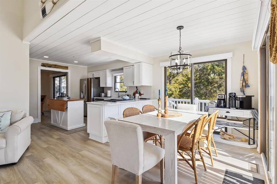 Gather around the sun-drenched dining table, where coastal charm meets modern comfort. Natural light pours through large windows, illuminating the open kitchen and creating your perfect space for memorable meals together.