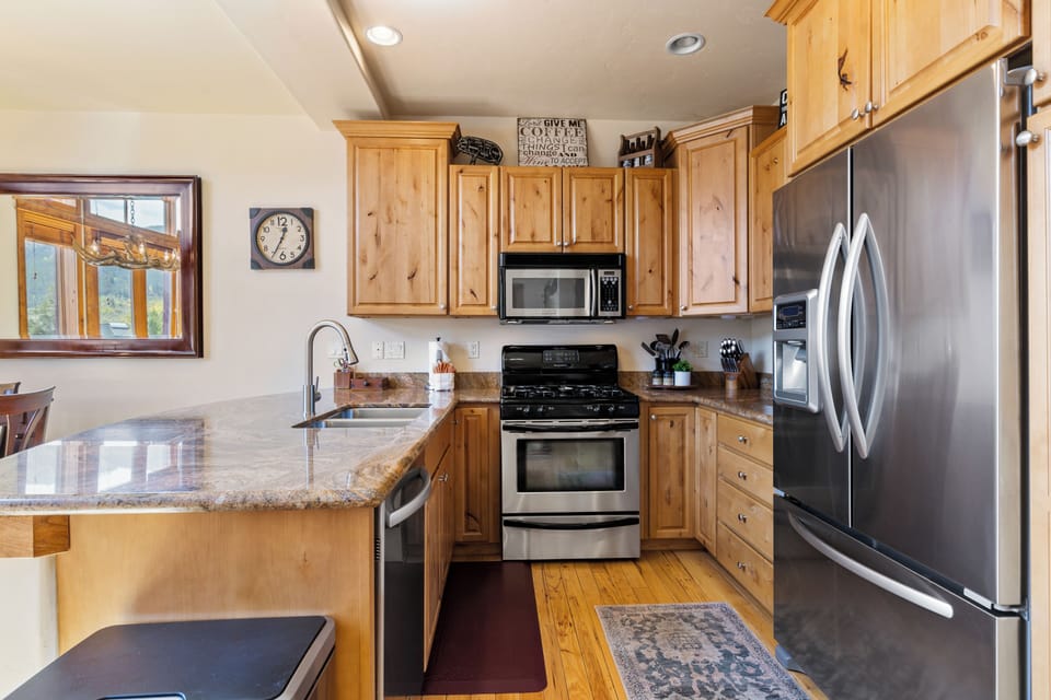 A modern kitchen with wooden cabinets, stainless steel appliances, and a marble countertop. A clock is on the wall above a window with a view.