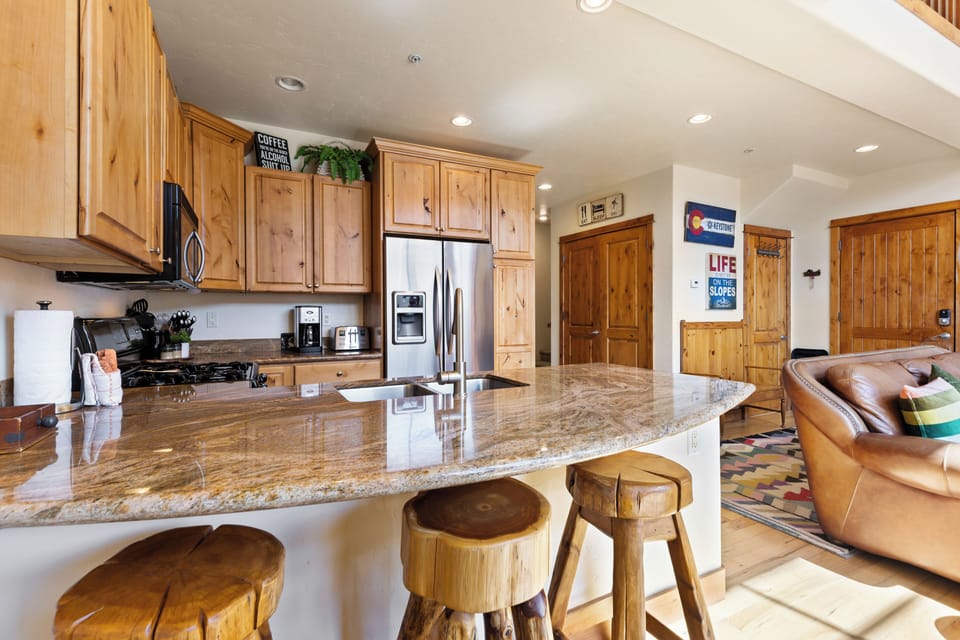 A kitchen with wooden cabinets, a granite countertop island with bar stools, and various appliances. A living area with a leather chair and rug is visible in the background.
