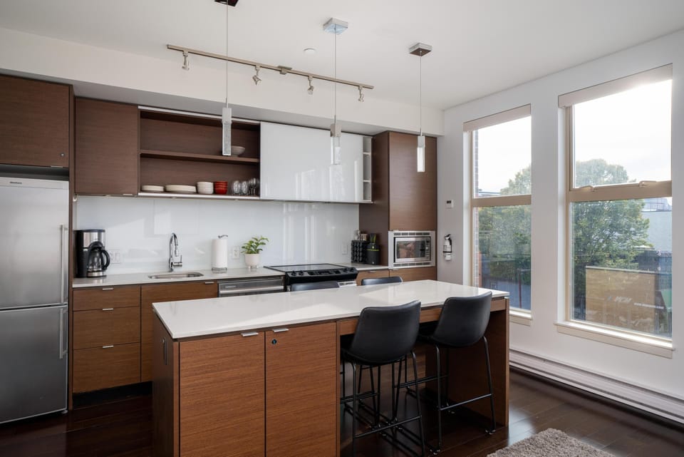 Broad angle connecting the kitchen galley, island prep surface, and natural light from tall exterior windows.