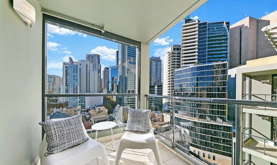 A glass-enclosed balcony space furnished with two white leisure chairs, overlooking surrounding high-rise buildings.